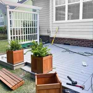 View of a partially constructed backyard deck with wooden planters and tools placed on a light gray deck surface, surrounded by grass and a lattice structure.