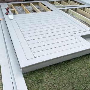 Close-up view of a partially completed floating deck with light gray composite decking boards laid out on a grassy area next to wooden framing.