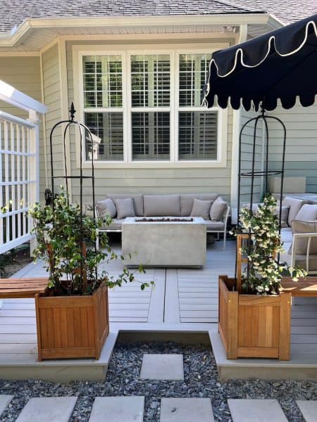 Cozy backyard patio featuring a comfortable sofa and a fire pit surrounded by wooden planters with climbing plants, set against a light-colored house.