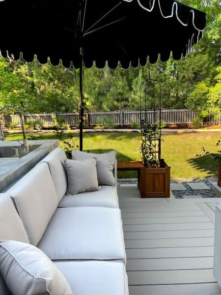 Cozy outdoor seating area featuring a white sofa with cushions, a large black umbrella, and greenery in the background.