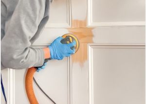A person in blue gloves sanding a wooden door panel using a power sander.