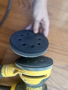 A hand holding a round sanding disc with holes, positioned above a yellow and black sander resting on wooden flooring.