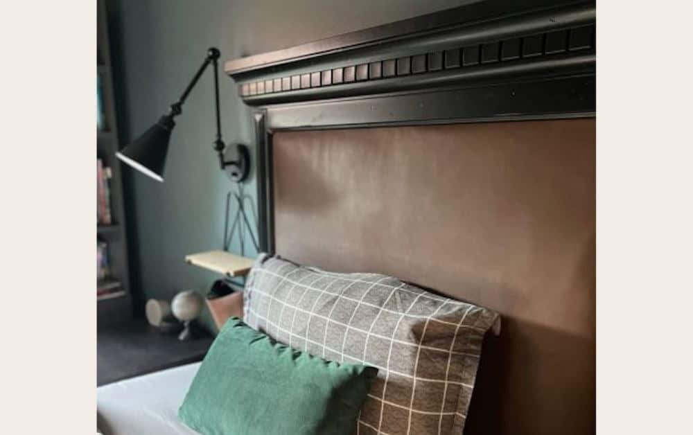A close-up view of a bed with a dark wooden headboard, featuring a brown upholstered panel, layered with a gray checkered pillow and a green accent pillow. A black wall lamp is mounted above the bed, and a bookshelf is visible in the background.