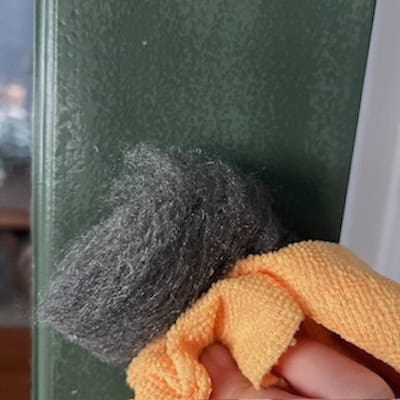 A person using a steel wool pad and an orange cloth to clean a green-painted surface.
