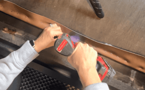 A close-up view of hands using a power drill to secure faux leather material to a backing board for a DIY headboard project.