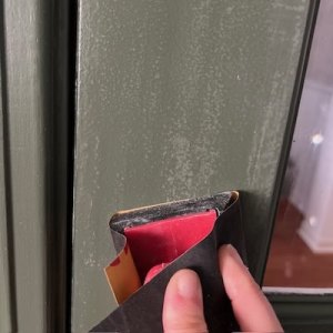 A hand holding a tool with a red and black protective cover, positioned near a door latch on a green door
