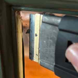 A close-up image of a person using a razor scraper to remove material from a surface, showing the blade and the edge of the surface being worked on.