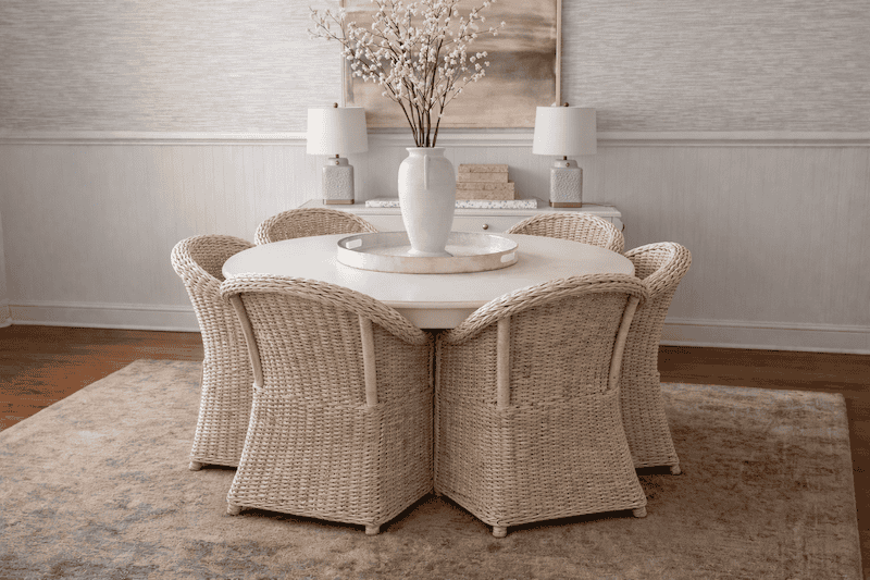 Neutral dining room decor featuring a round white table, four woven wicker dining chairs, and a styled console with centered artwork and lamps.