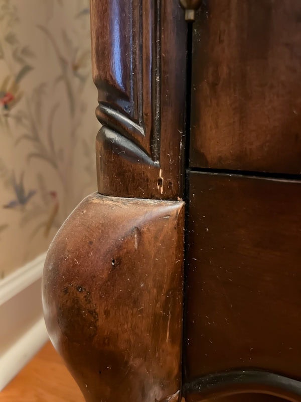 Close-up of a wooden furniture corner showing detailed craftsmanship and wear, set against a floral wallpaper background.