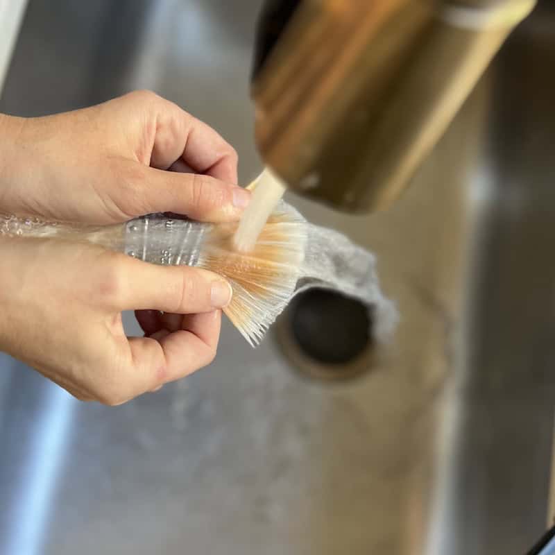 A person washing a brush under running water in a sink.