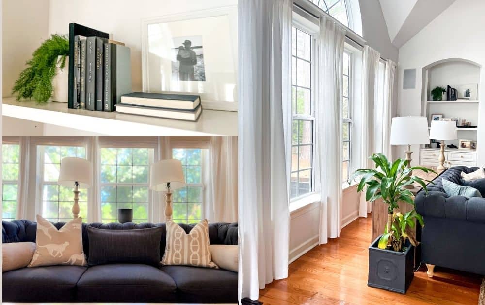 A cozy living room featuring a black sofa with decorative pillows, a large window letting in natural light, a plant in a corner, and books on a shelf.