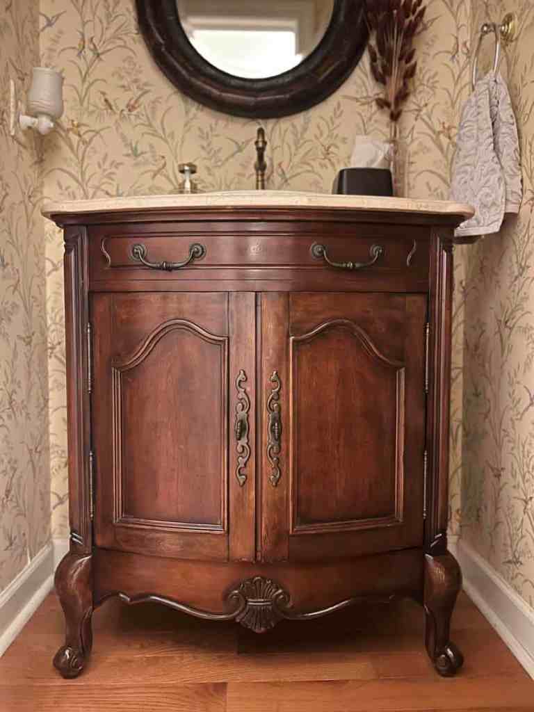 A wooden vanity cabinet with two doors and decorative hardware, situated in a bathroom with floral wallpaper and a round mirror in the background.