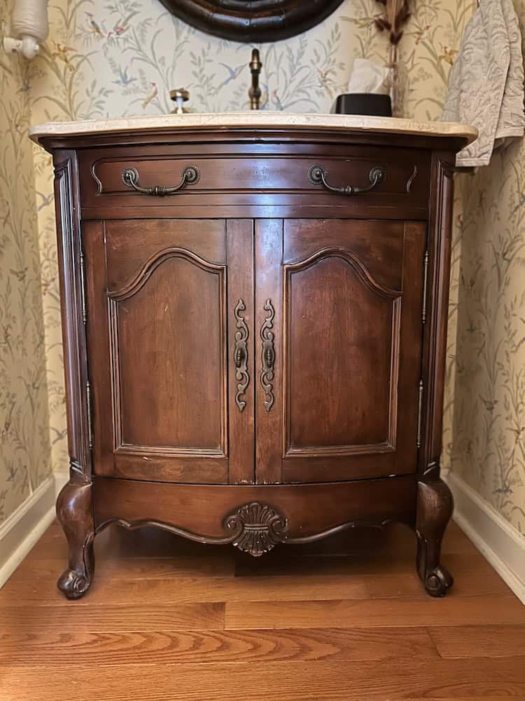 A vintage wooden cabinet with ornate detailing, featuring two doors and decorative hardware, set against a floral wallpaper background.
