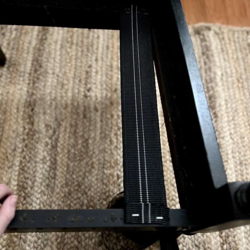 Close-up of a black strap laid across the underside of a wooden chair frame, with a hand holding the frame. The background features a woven rug.