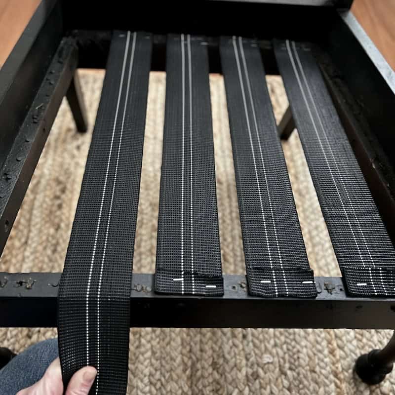 Close-up of a black bed frame showing woven slats with white stitching, with a hand adjusting one of the slats.