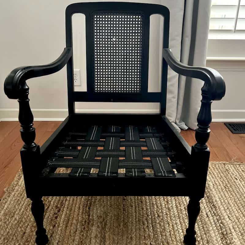 A black wooden chair with a caned backrest and a woven seat frame, positioned on a natural fiber rug.