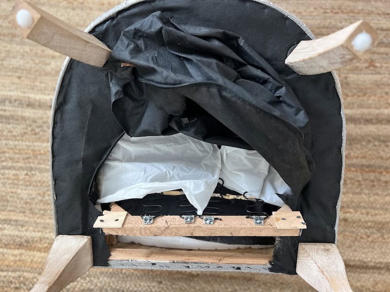 Top view of the underside of a furniture piece, showing wooden legs, black fabric, and exposed components including springs and padding.