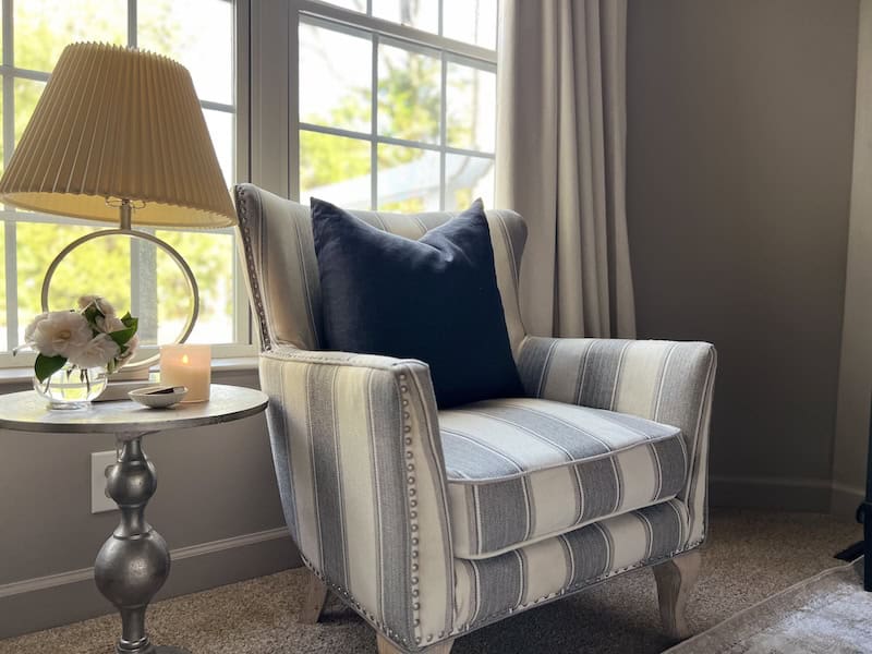 A cozy, striped armchair with a dark blue pillow, next to a side table featuring a lamp, a small vase of flowers, and a lit candle, in a bright room with large windows.