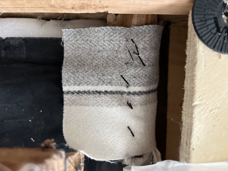 A close-up view of a piece of gray and white fabric being sewn, with visible black stitches and pins securing it to a wooden structure.