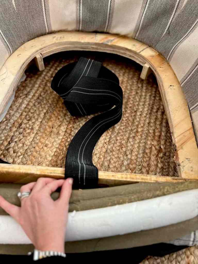 A hand holding a black strap inside a furniture frame made of wood, with woven material below and a striped fabric visible in the background.