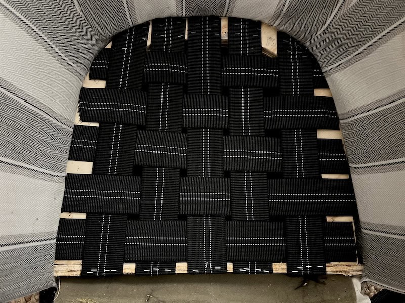 A close-up view of a woven black seat made from patterned straps, placed in a wooden frame.