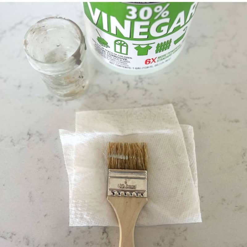 A bottle of 30% vinegar next to a glass jar and a paintbrush resting on a paper towel, all placed on a marble surface.