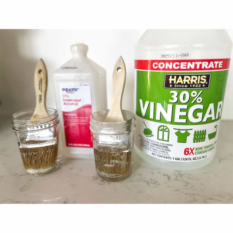 Two jars with wooden brushes soaking in liquids, next to a bottle of isopropyl alcohol and a large container of 30% vinegar on a countertop.