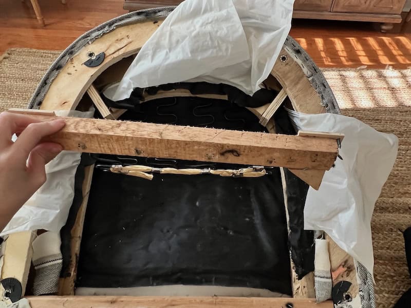 A close-up image showing a person's hand holding a wooden piece while examining the inside of a chair, which is partially disassembled. The chair's frame is visible with black material and some white fabric covering it.