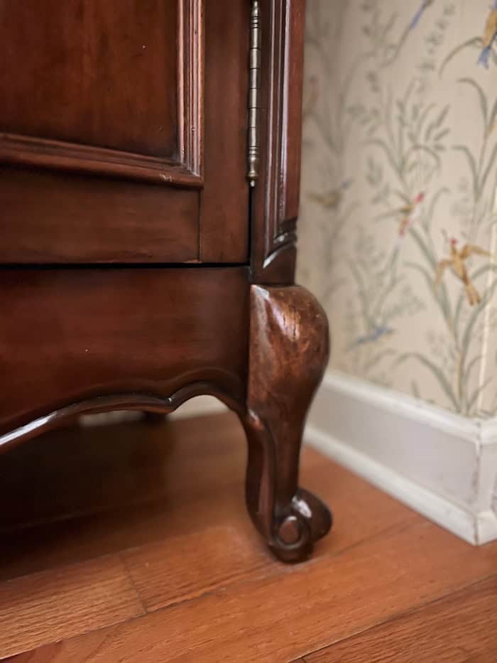 Close-up of the wooden leg of a furniture piece featuring a decorative design, with a floral wallpaper in the background.