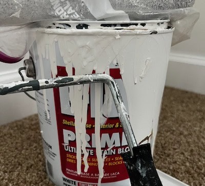 Close-up of a paint bucket with dripping white paint and a paint roller resting on the side.