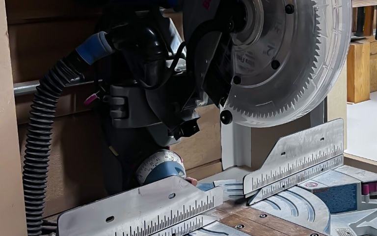 A close-up view of a Bosch sliding compound miter saw in a workshop, showing the circular blade, protective guard, and metal fence used for precise angled cuts in DIY home renovations.