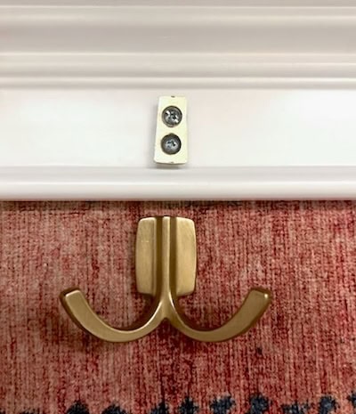 A close-up of a gold double hook mounted on a white wall, with a wall plate and screws visible above it, on a textured red rug.