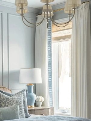 A serene bedroom featuring light blue walls, white drapery, and a window with natural light. A bedside table holds a blue lamp and a small vase of white flowers, complemented by elegant bedding.