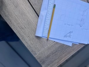 Hand-drawn sketches on graph paper showing dimensions for a DIY wood project — including a box frame labeled '20 inches long, 2x4' and '17.5 inches height,' with 45-degree angle cuts noted. A yellow pencil and ruler rest on top of the papers, set on a weathered gray wood surface.