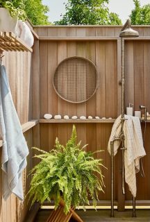Warm wood outdoor shower enclosure with built-in shelf, round mirror, towels and potted fern