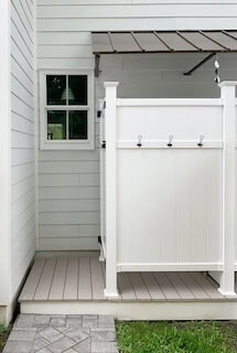 Outdoor white wooden shower stall with four hooks on the side, next to a window on light gray siding wall